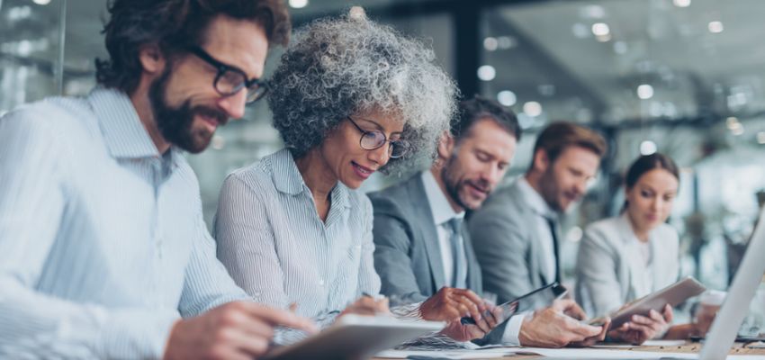 Diverse team of business professionals in a modern office collaboratively examining documents and data on tablets during a group contract auditing meeting.